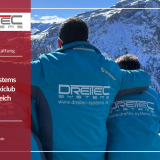 Drei Männer in blauen Skijacken mit dem Rücken zur Kamera und dem DreiTec Systems-Logo auf dem Rücken schauen von einer Holzterrasse auf eine sonnige und verschneite Skipiste, im Hintergrund die Berge Österreichs